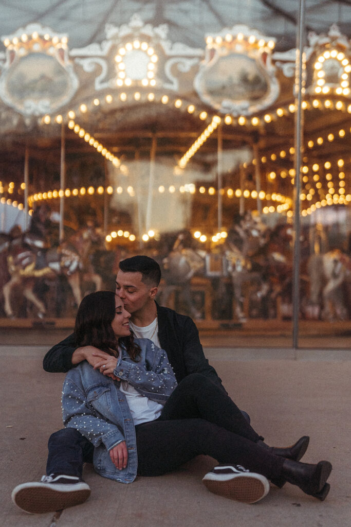 Man kissing woman's forehead during their Brooklyn engagement session with a carousel in the background.