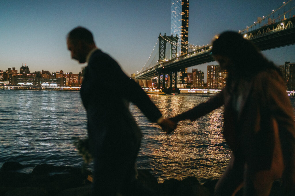 Couple holding hands with the Brooklyn Bridge in the background at dusk.
