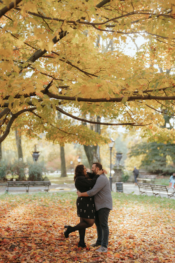 Couple hugging in Prospect Park in NYC in the fall with colorful fall trees surrounding them. 