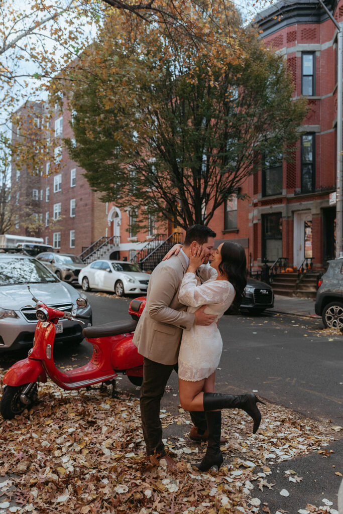 Engaged couple embracing beside a red scooter on an autumn Brooklyn street covered in fall leaves.