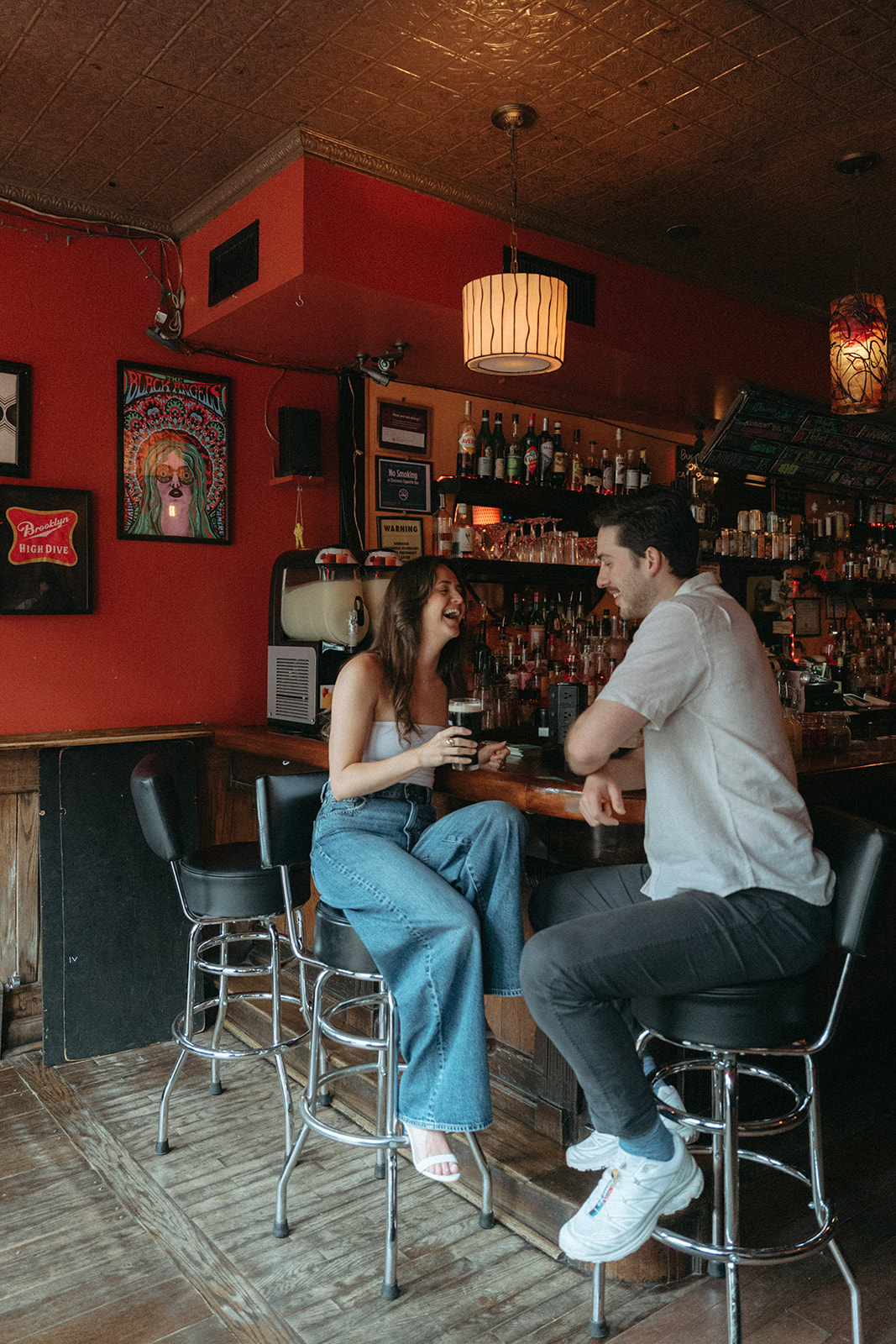 Engaged couple sitting on stools at a red-walled Brooklyn bar during their engagement session.
