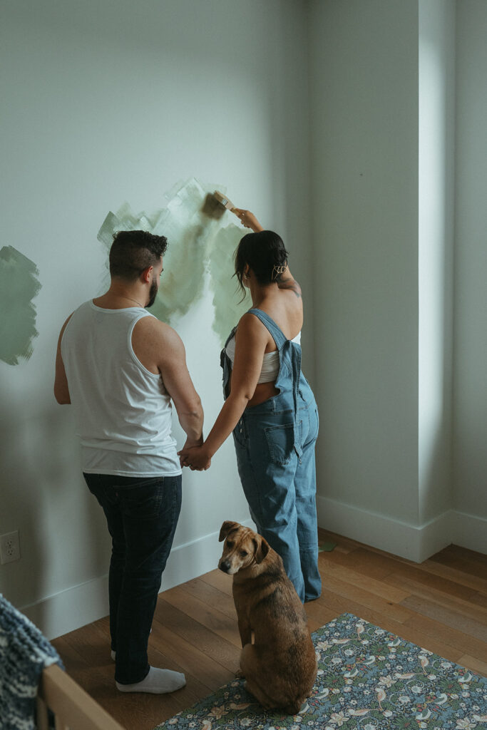 Maternity photography where husband and pregnant wife paint the nursery for their baby-to-be. Their dog is watching them. 