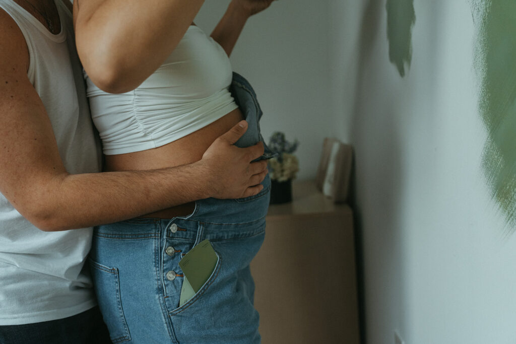 A close up photo of a husband holding his wife's pregnant belly while they paint the nursery together for their at home maternity photography session.