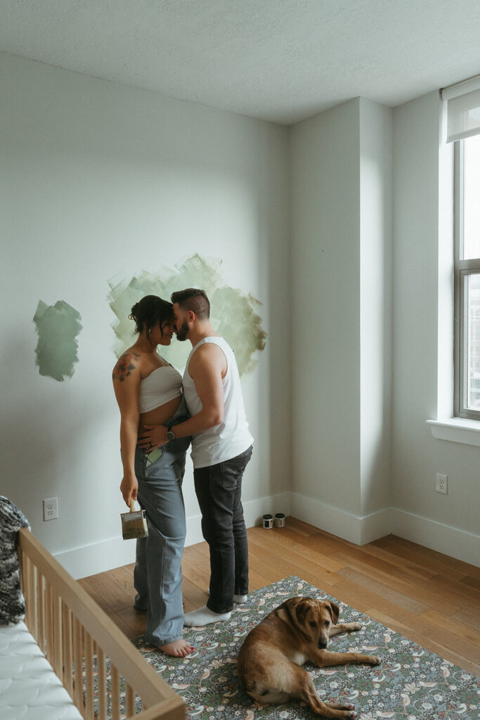 A husband and pregnant wife paint the nursery for their baby-to-be. Their dog is watching them. 