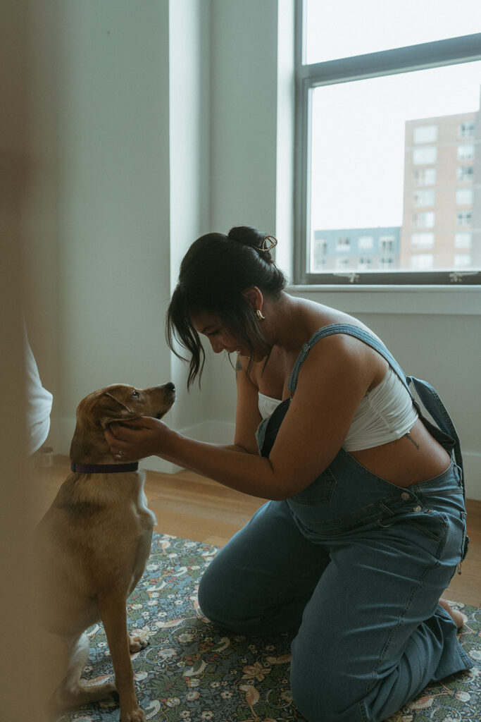 A pregnant woman wearing overalls petting her dog.