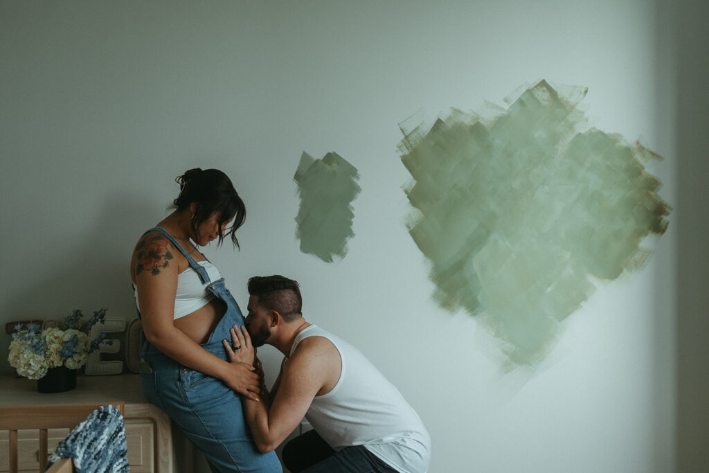 A husband and pregnant wife are painting the nursery for their baby-to-be, and the husband stops to kiss his wife's belly.