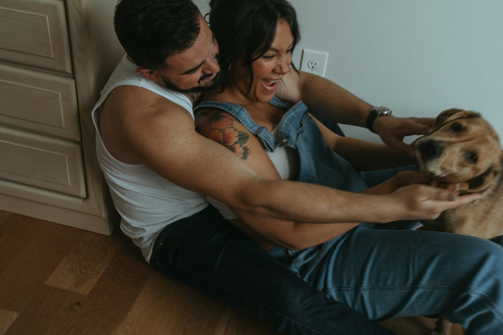 A husband and pregnant wife are cuddling on the floor of their nursery with their dog.