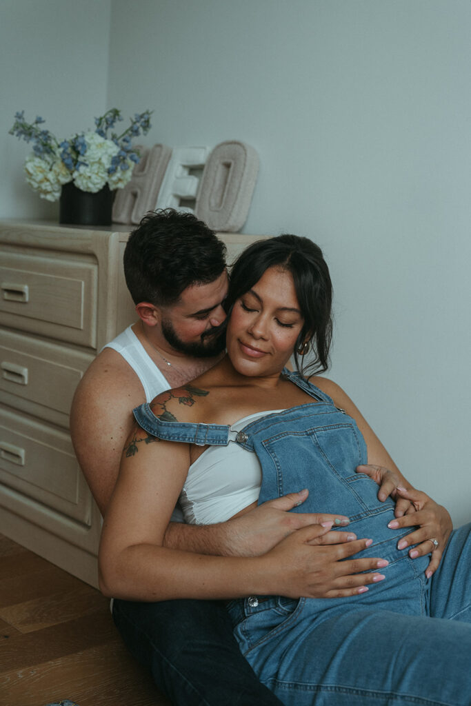 A husband and pregnant wife sit on the floor of the nursery for their baby-to-be, intimately cuddling for an at home maternity photography session in NYC.