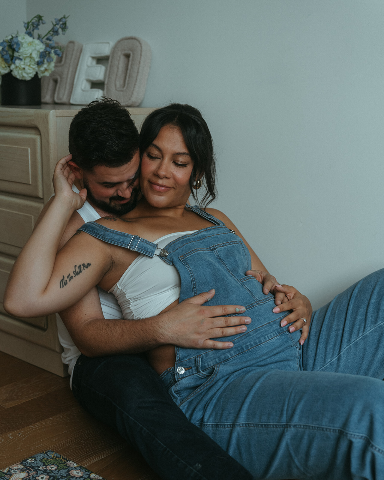 A husband and pregnant wife sit on the floor of the nursery for their baby-to-be, intimately cuddling.