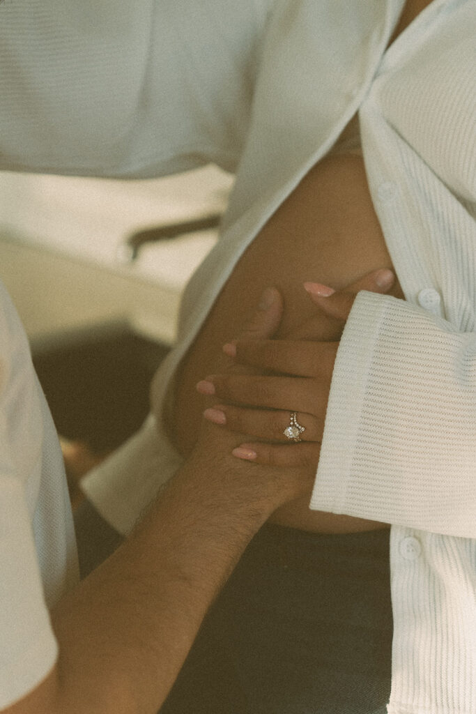 A close up photo of a husband holding his pregnant wife's belly, featuring the wife's wedding rings for a maternity photography session.