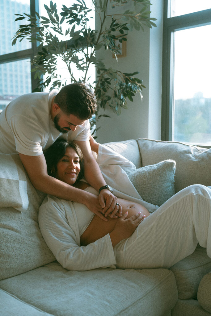 A husband and pregnant wife cuddling together on the couch in their NYC apartment for a maternity photography session.