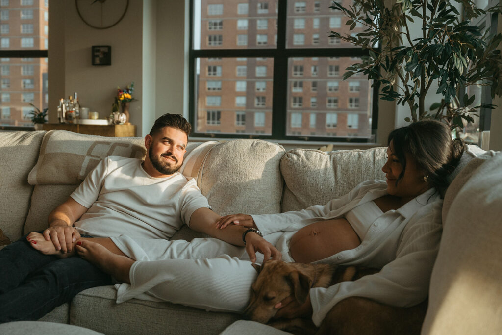 A husband and pregnant wife cuddling together with their dog on the couch in their NYC apartment for a maternity photography session.