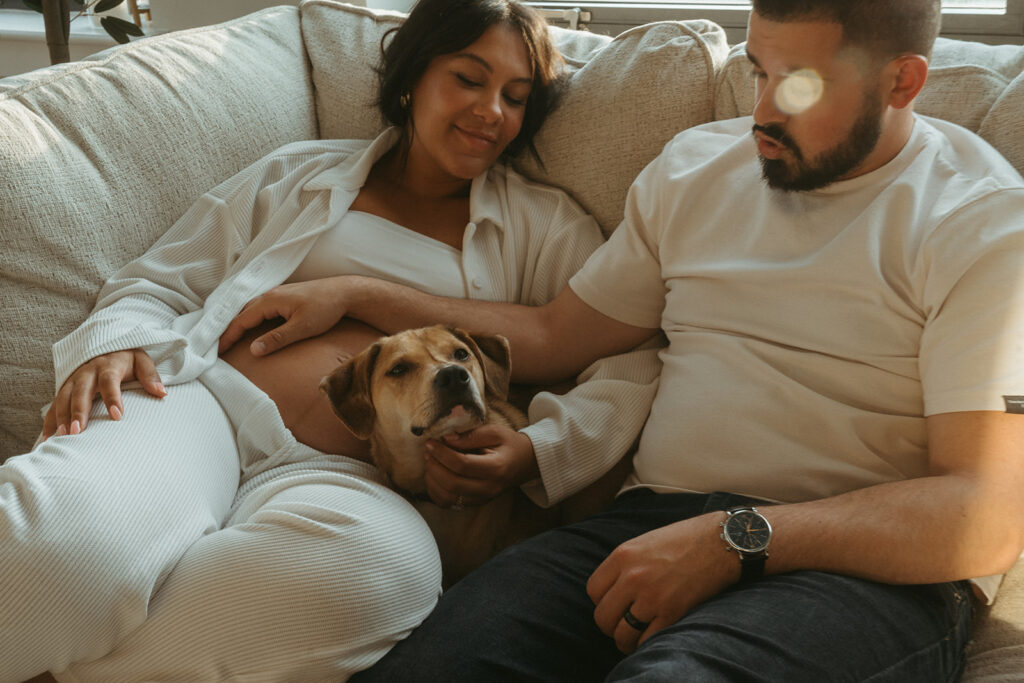 A husband and pregnant wife cuddling together with their dog on the couch in their NYC apartment.