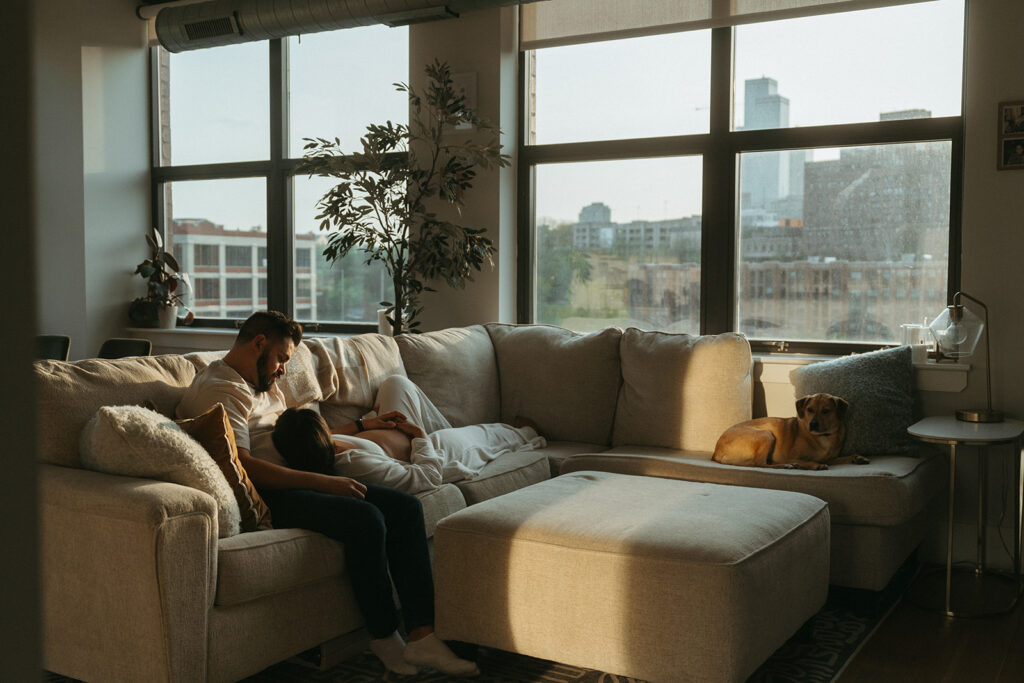 A husband and pregnant wife cuddling together on the couch in their NYC apartment.