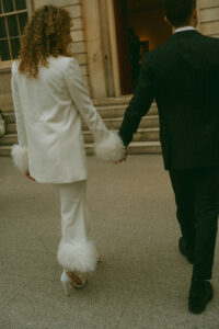 Bride in a white feather-cuffed pantsuit walking off with her groom after eloping in NYC