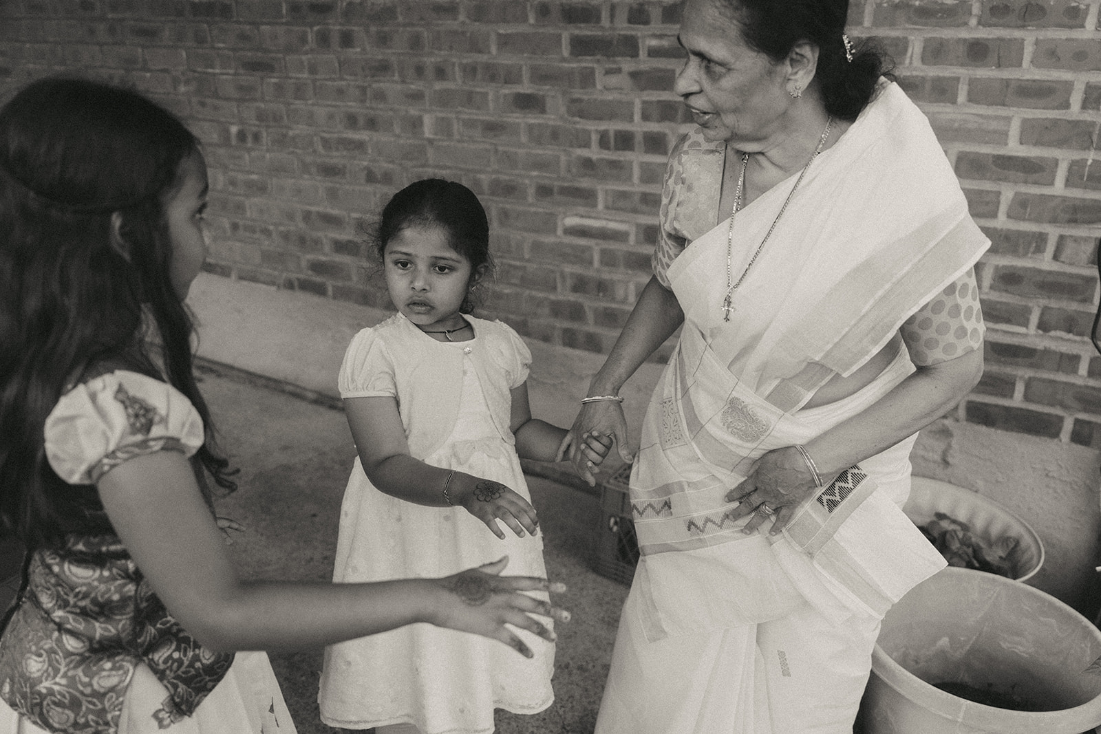 Alt Text: A grandmother gently holding the hands of two young girls during a quiet moment at a Catholic Indian wedding, highlighting cross-generational connection and tradition, captured in black and white by Elsie Goodman Photography at an Indian wedding venue on Long Island.