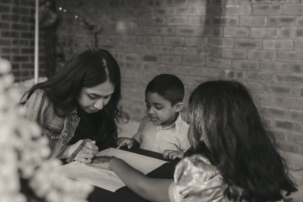 Alt Text: A woman applying intricate mehndi to a young girl’s hand while a young boy watches curiously, showcasing pre-wedding rituals at a Catholic Indian wedding at a Long Island wedding venue, beautifully documented by Elsie Goodman Photography.
