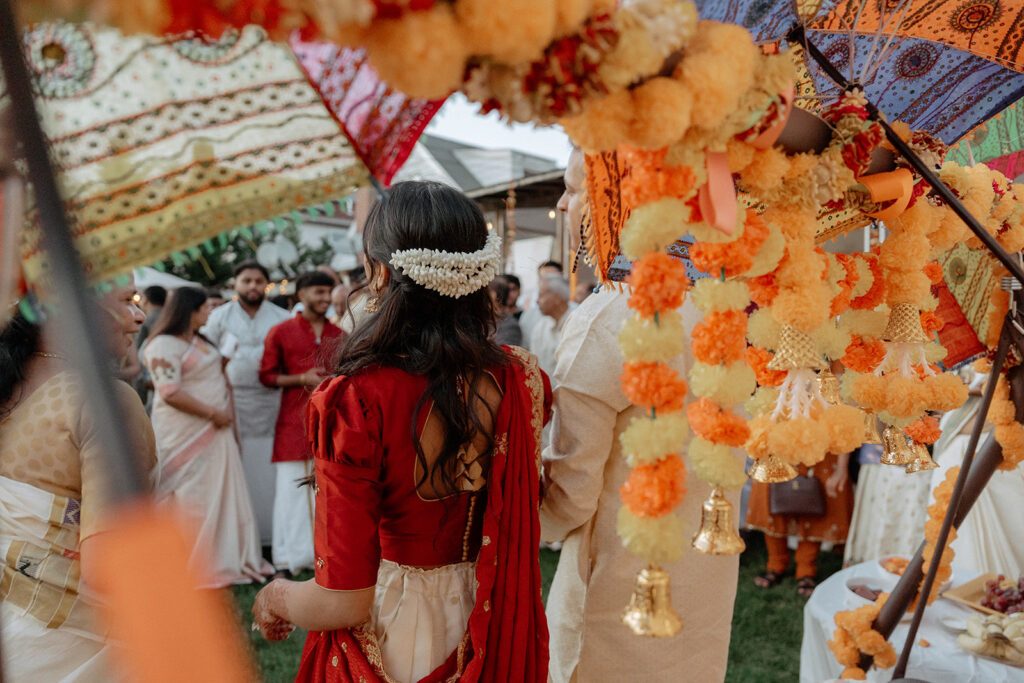 Alt Text: The bride seen from behind beneath vibrant floral umbrellas and garlands during a joyful outdoor celebration at an Indian Catholic wedding on Long Island, framed by traditional decorations and guests in festive attire, photographed by Elsie Goodman Photography.