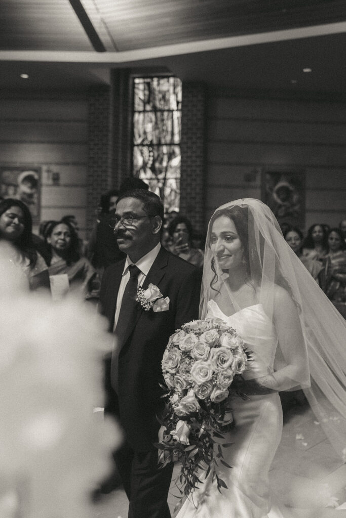 The bride walks down the aisle holding her father’s arm, smiling at the groom during a heartfelt Catholic Indian wedding ceremony at a Long Island church, beautifully captured by Elsie Goodman Photography for an Indian wedding photo gallery.
