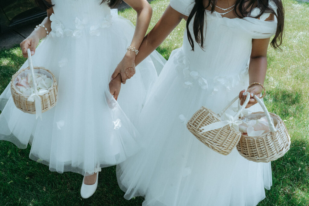 Two young flower girls in matching white dresses hold hands and carry baskets of petals outside a Catholic Indian wedding ceremony, a sweet candid moment captured by Elsie Goodman Photography for an Indian wedding photo gallery.