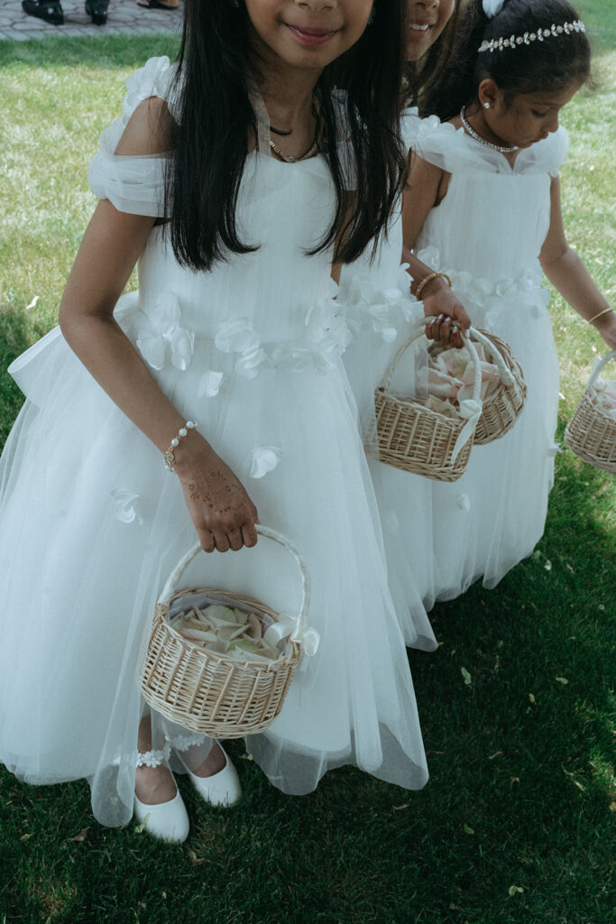 Flower girls in white dresses holding woven baskets of rose petals stand together outside during a Catholic Indian wedding celebration, captured beautifully by Elsie Goodman Photography for an Indian wedding photo gallery.