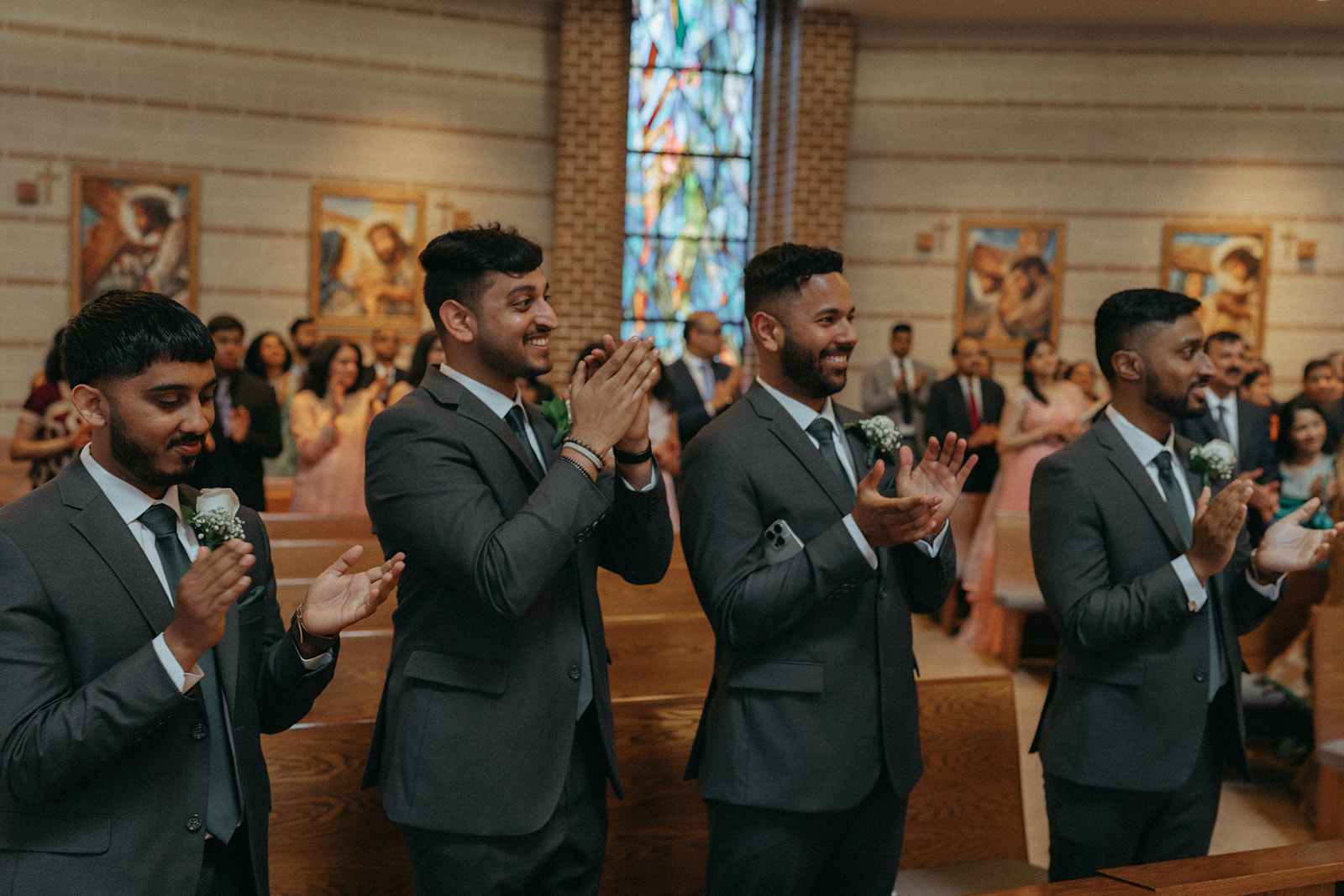 The groomsmen clap and smile during the Catholic Indian wedding ceremony, celebrating the newlyweds in a Long Island church, a joyful candid moment captured by Elsie Goodman Photography for an Indian wedding photo gallery.