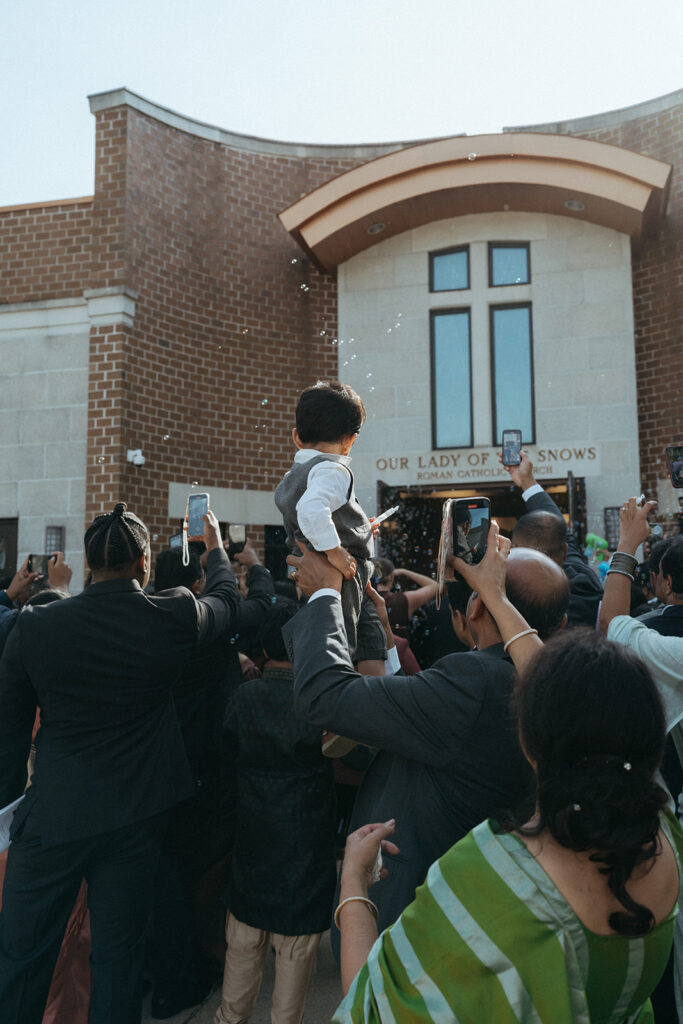 A child is lifted above the crowd outside Our Lady of the Snows Roman Catholic Church, with bubbles filling the air to celebrate a Catholic Indian wedding exit, a lively moment captured by Elsie Goodman Photography for an Indian wedding photo gallery.