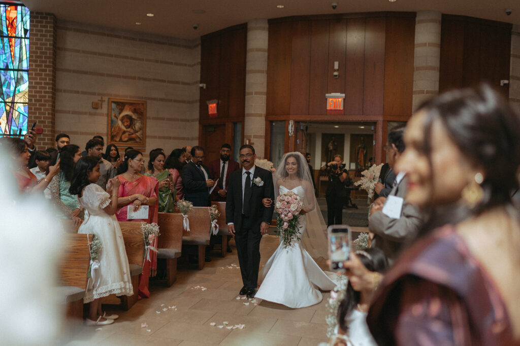 The bride walks down the aisle holding her father’s arm, smiling at the groom during a heartfelt Catholic Indian wedding ceremony at a Long Island church, beautifully captured by Elsie Goodman Photography for an Indian wedding photo gallery.