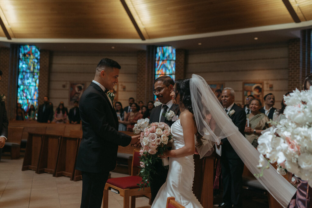 The bride, escorted by her father, smiles at the groom during their first meeting at the altar in a Long Island Catholic Indian wedding, a heartfelt moment documented by Elsie Goodman Photography for an Indian wedding photo gallery.