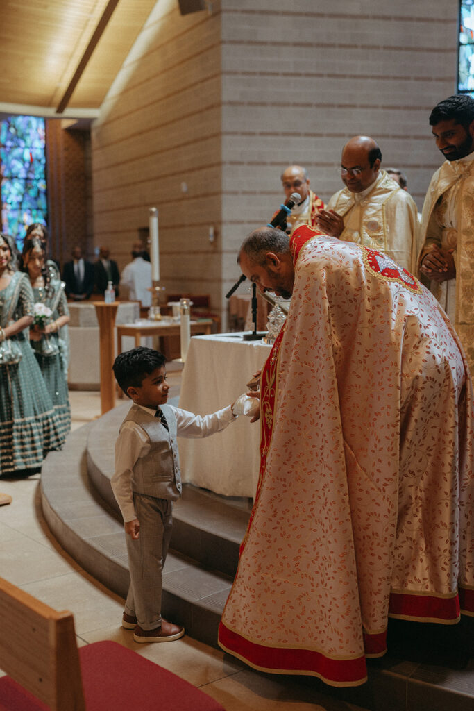 A young boy in a gray vest and tie offers rings to a priest during a Catholic Indian wedding ceremony at a Long Island church, beautifully captured by Elsie Goodman Photography for an Indian wedding photo gallery.