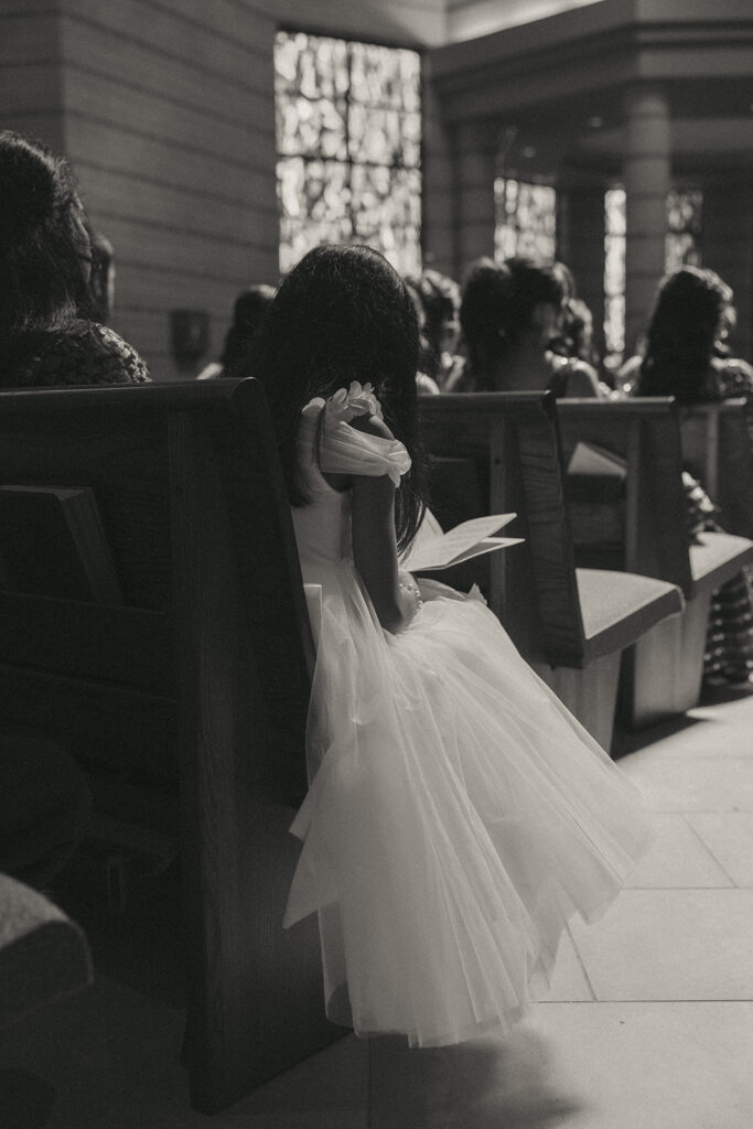 A young flower girl in a white tulle dress sits quietly in a pew, holding her ceremony program during a Catholic Indian wedding, an intimate moment captured by Elsie Goodman Photography for an Indian wedding photo gallery.