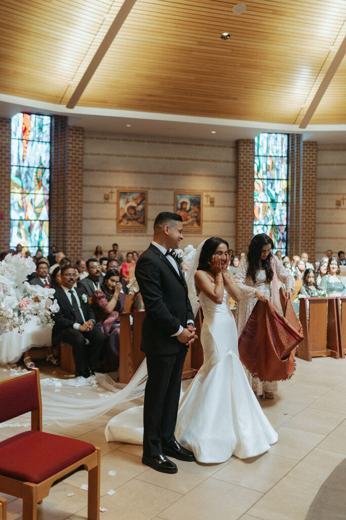 The bride clasps her hands to her face in joyful emotion during the Catholic Indian wedding ceremony while the groom smiles beside her, a candid moment captured by Elsie Goodman Photography for an Indian wedding photo gallery.