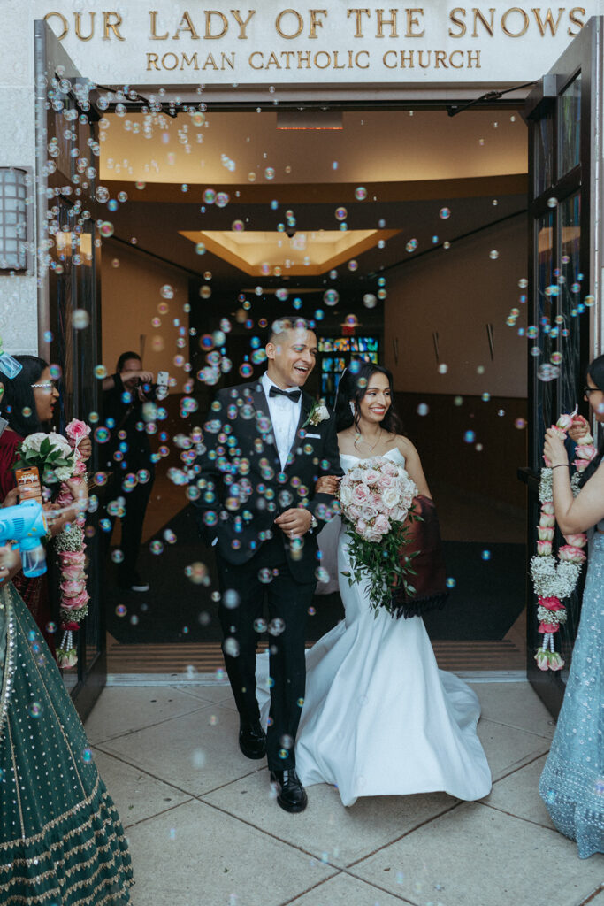 Joyful bubble exit at Our Lady of the Snows Roman Catholic Church as the bride and groom smile and walk hand in hand during their Long Island Indian Catholic wedding, photographed by Elsie Goodman Photography.
