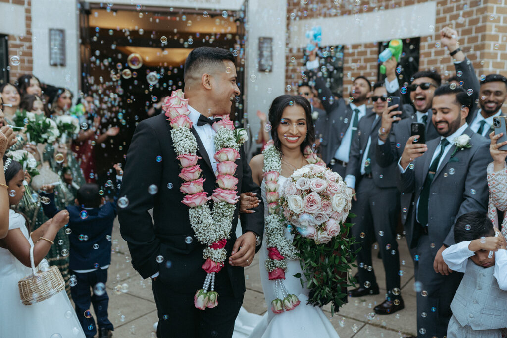 Newlywed couple smiles while walking out of Our Lady of the Snows Roman Catholic Church, surrounded by bubbles and cheering guests, during their Catholic Indian wedding captured by Elsie Goodman Photography.