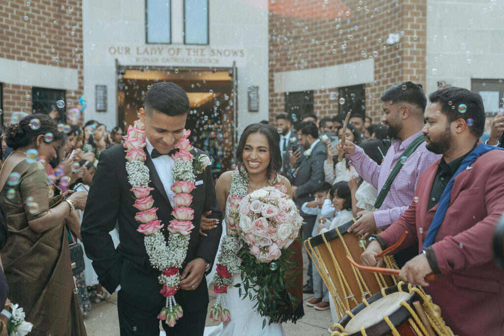 Joyful bubble exit at Our Lady of the Snows Roman Catholic Church as the bride and groom smile and walk hand in hand during their Long Island Indian Catholic wedding, photographed by Elsie Goodman Photography.