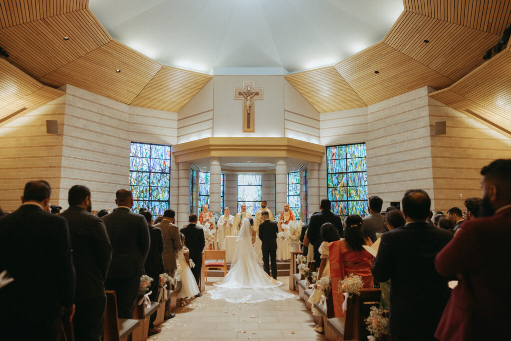 The bride and groom stand before the altar surrounded by family and friends during a Catholic Indian wedding mass at Our Lady of the Snows Church, a stunning moment captured by Elsie Goodman Photography for an Indian wedding photo gallery.