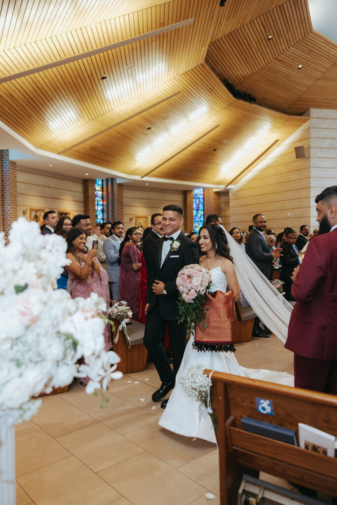 The bride and groom walk down the aisle smiling after their Indian Catholic wedding ceremony at Our Lady of the Snows, with guests cheering and photographing the moment, captured by Elsie Goodman Photography.