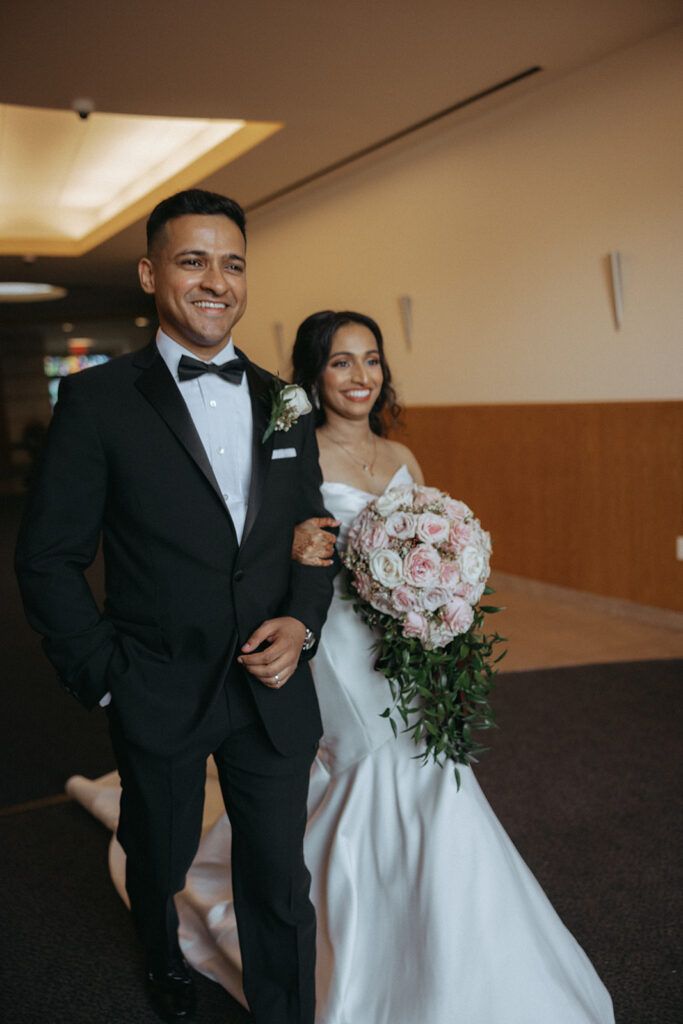 Smiling bride and groom walk together down the church hallway during their Long Island wedding day, the bride carrying a cascading bouquet of pink roses, captured by Elsie Goodman Photography.
