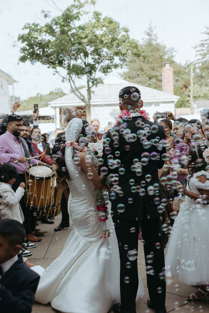 Back view of the bride and groom walking through a joyful bubble exit, with guests celebrating their Indian Catholic wedding ceremony at Our Lady of the Snows, captured by Elsie Goodman Photography.