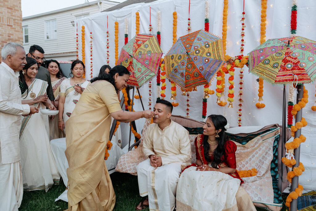 Mother feeding sweets to groom while bride smiles during Catholic Indian wedding celebration — Elsie Goodman Photography.
