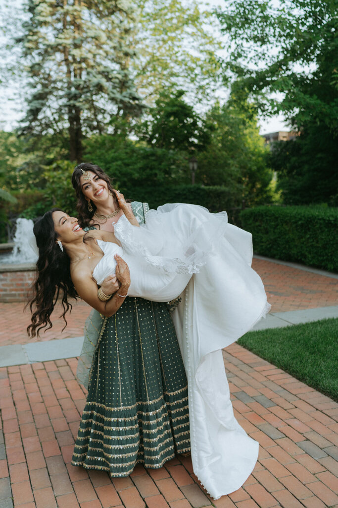 Bridesmaid lifts the bride playfully, both laughing in a joyful outdoor moment showcasing Indian Catholic wedding dresses, photographed by Elsie Goodman Photography.