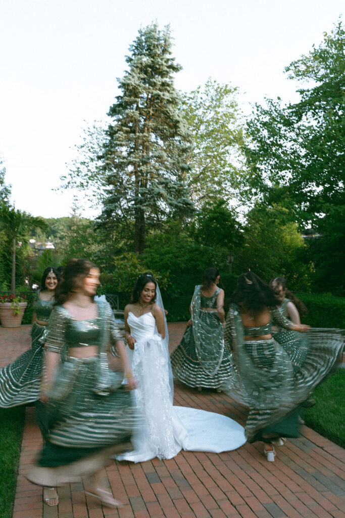 Bride laughing as bridesmaids spin in motion around her in green and gold lehengas during wedding party photos by Elsie Goodman Photography.