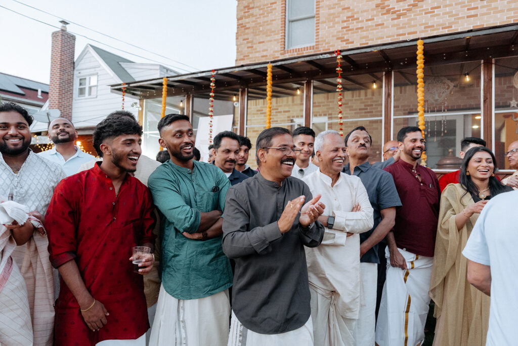 A joyful group of guests dressed in traditional Indian attire, including kurtas and veshtis, laughing and clapping during an outdoor night before Catholic Indian wedding in Long Island, captured by Elsie Goodman Photography.