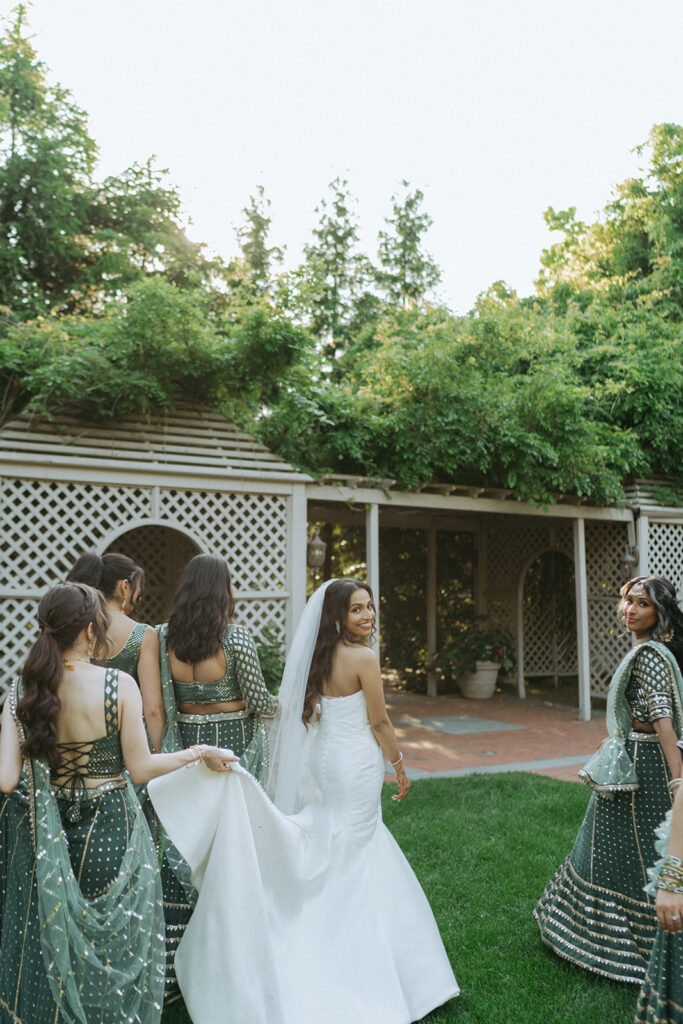 Bride smiling back at the camera while bridesmaids adjust her train, a beautiful Indian Catholic wedding party photo by Elsie Goodman Photography.