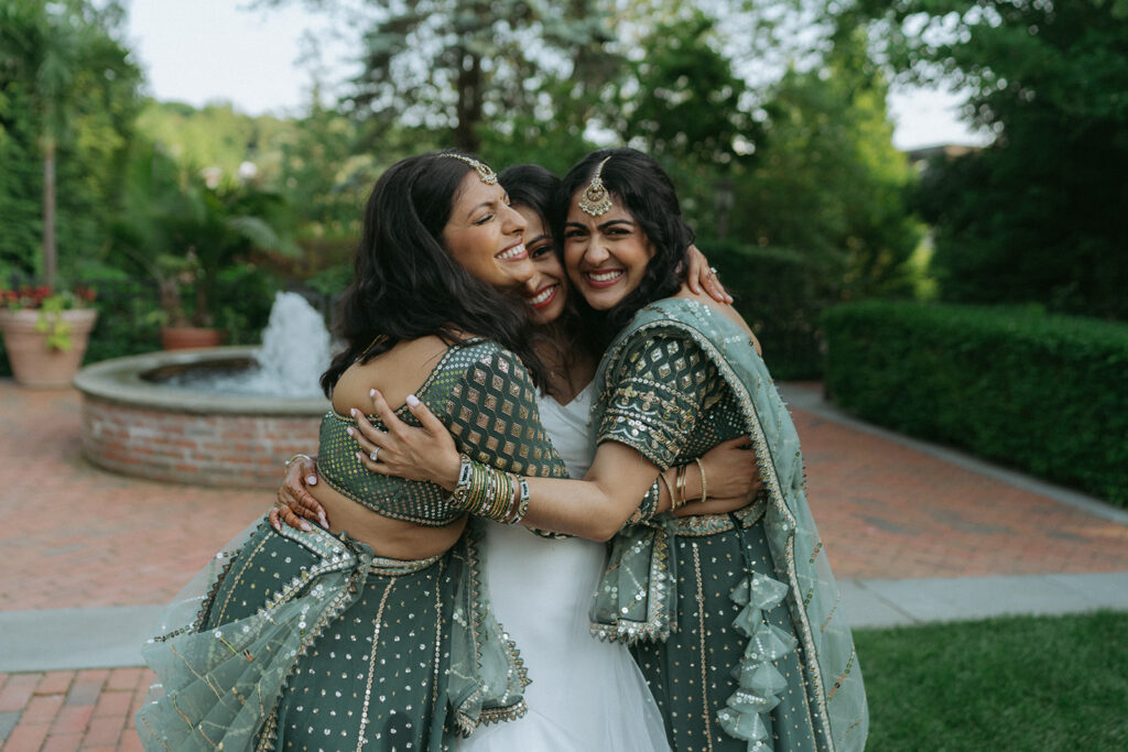 Bride hugging two bridesmaids near a fountain, all beaming with happiness in colorful Indian Catholic wedding dresses, photographed by Elsie Goodman Photography.