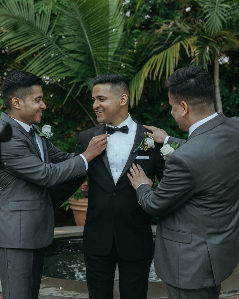 Groom getting his bow tie adjusted by groomsmen in a joyful outdoor moment, photographed by Elsie Goodman Photography at an Indian Catholic wedding.