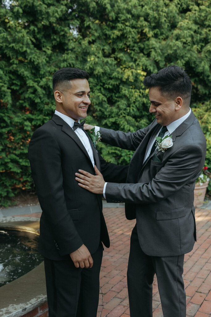 Groom smiling as his groomsman helps adjust his boutonnière before the ceremony, captured by Elsie Goodman Photography during an Indian Catholic wedding.