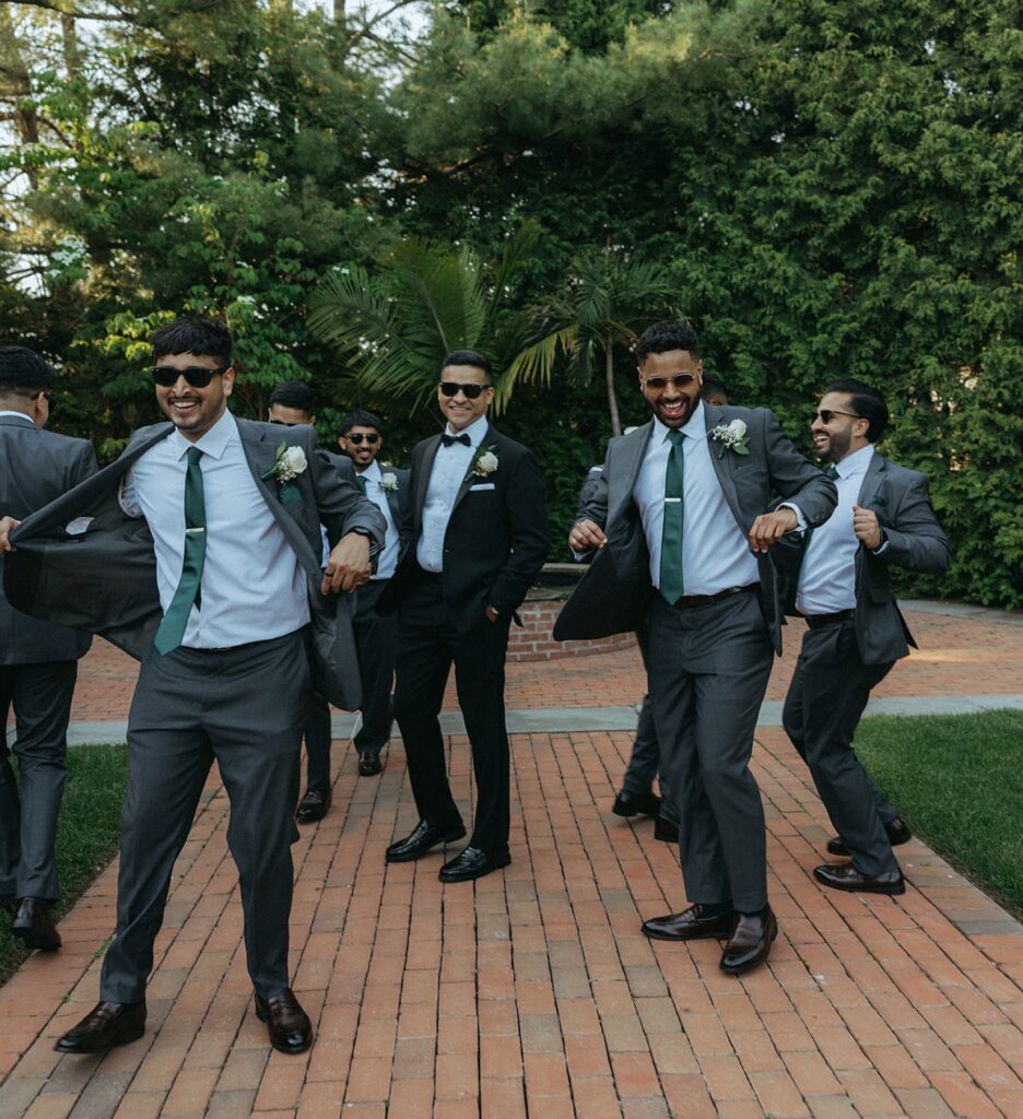 The groom laughing with his groomsmen in stylish gray suits and green ties during a fun outdoor portrait session at a Catholic Indian wedding at Crest Hollow Country Club, captured by Elsie Goodman Photography at one of the top Indian wedding venues on Long Island.