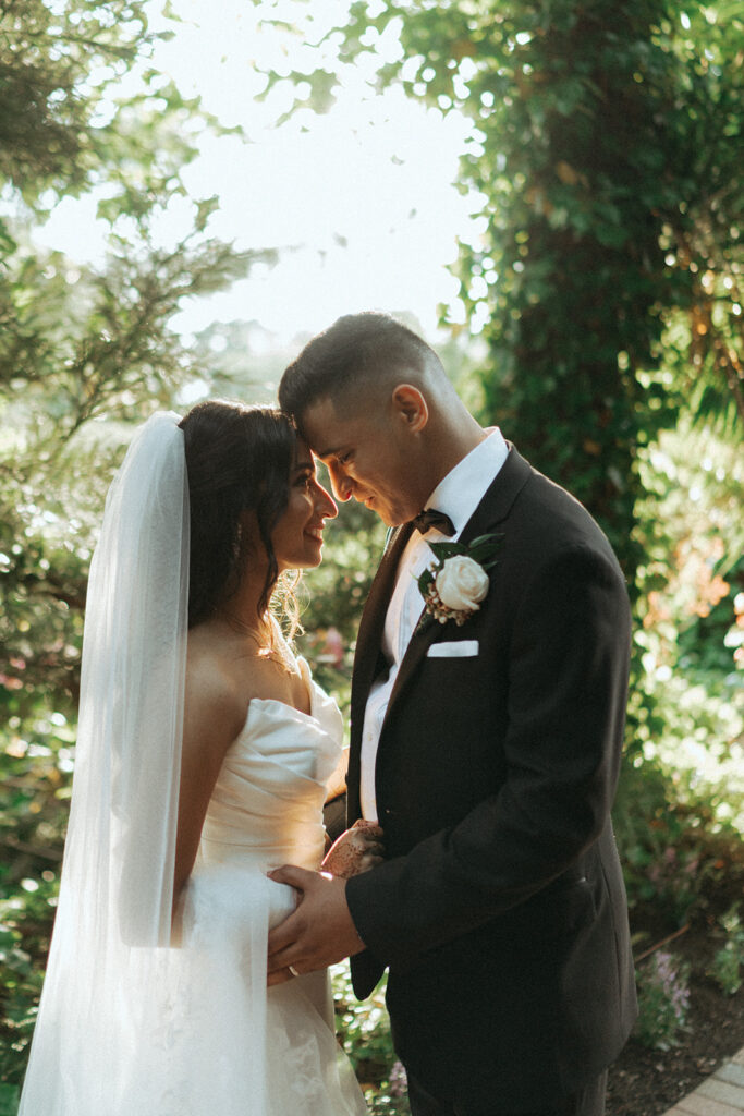 The bride and groom smile with their foreheads touching, surrounded by lush gardens during their Catholic Indian wedding at Crest Hollow Country Club, captured by Elsie Goodman Photography.