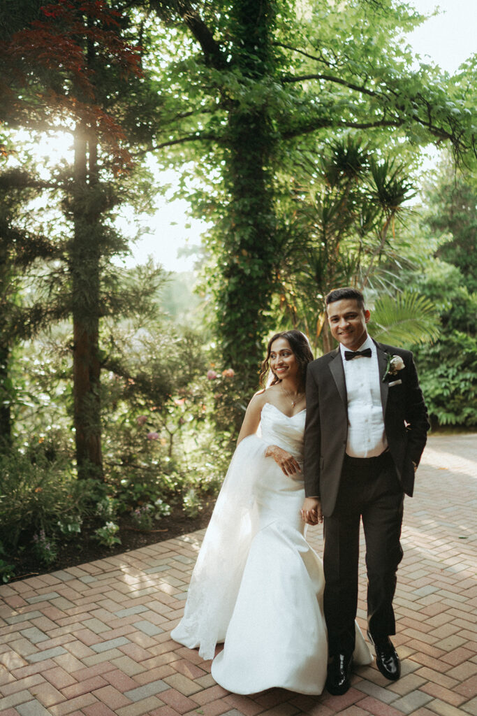 The bride and groom walk hand in hand through lush gardens at their Crest Hollow Country Club wedding, a beautiful Indian wedding venue in Long Island, captured by Elsie Goodman Photography.